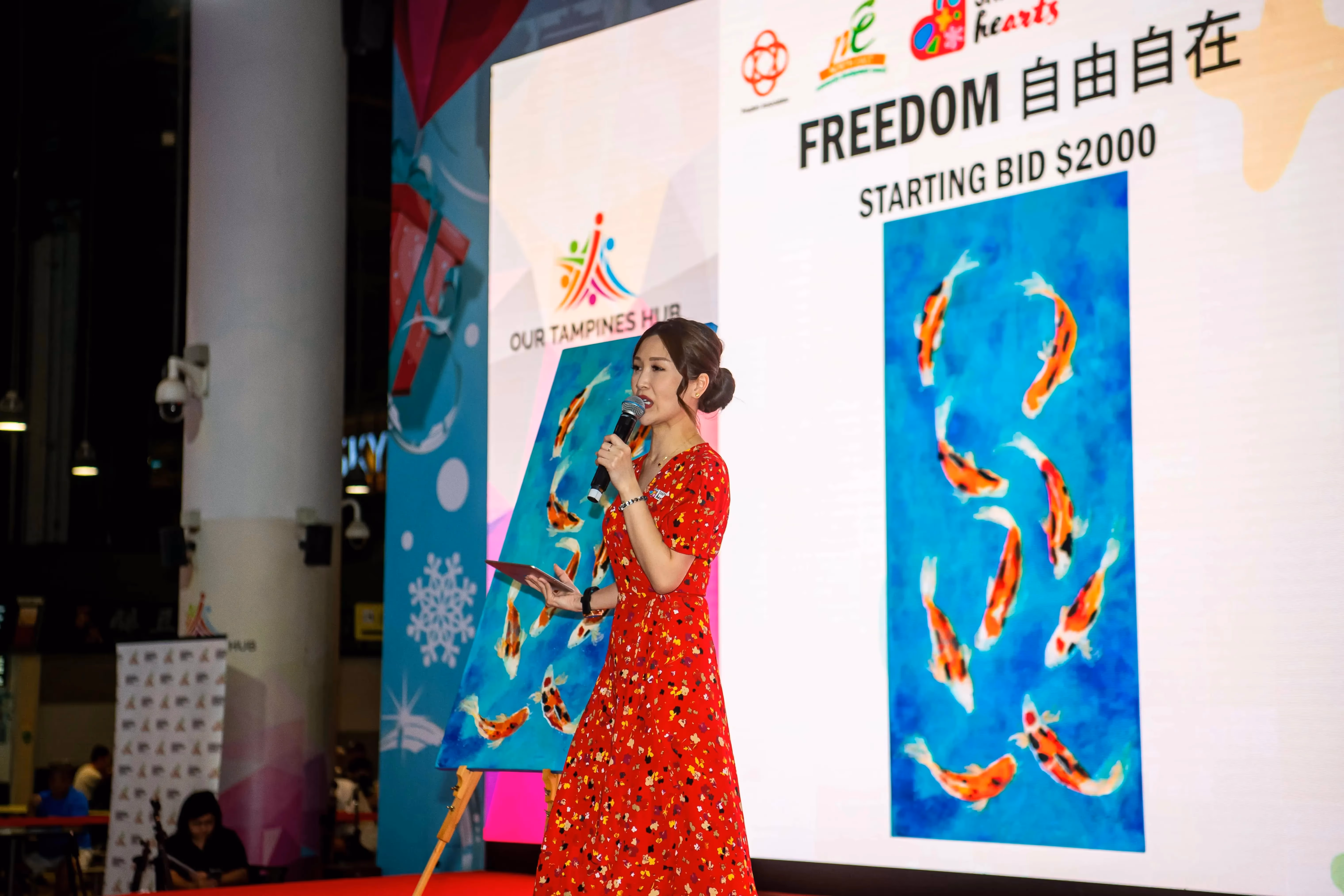 A person in a red dress is presenting a painting of koi fish with a starting bid of $2000 at Our Tampines Hub.